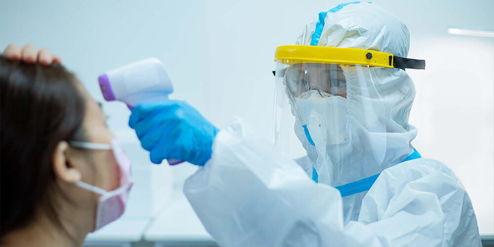 A doctor wearing full PPE holding a thermometer to a patients head